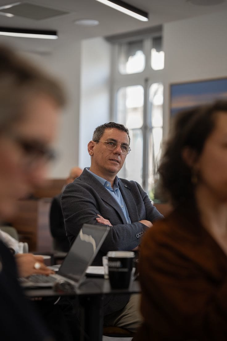 Empresário de óculos e braços cruzados escuta com atenção numa sala de reunião, com outras pessoas desfocadas em primeiro plano